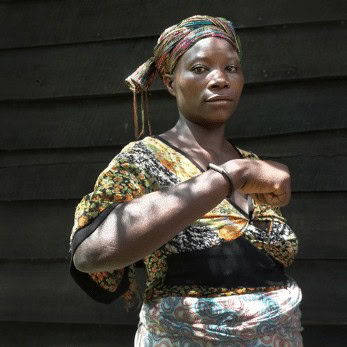 A woman with traditional clothing and a colorful headscarf standing against a dark wooden wall.