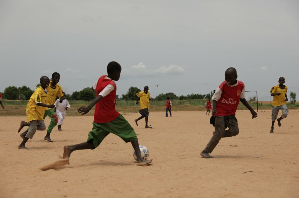 Children playing soccer on a dusty field, enjoying outdoor activity in a rural setting, wearing colorful sports jerseys under an overcast sky.