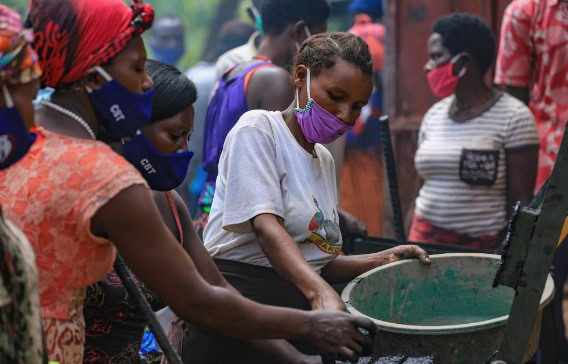 Women wearing face masks distribute water from a large container to community members during a humanitarian aid effort.