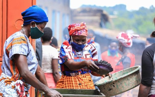 A woman preparing food over an open flame in a busy outdoor setting, with other community members in the background. The scene highlights efforts to support local livelihoods in a crisis-affected area.