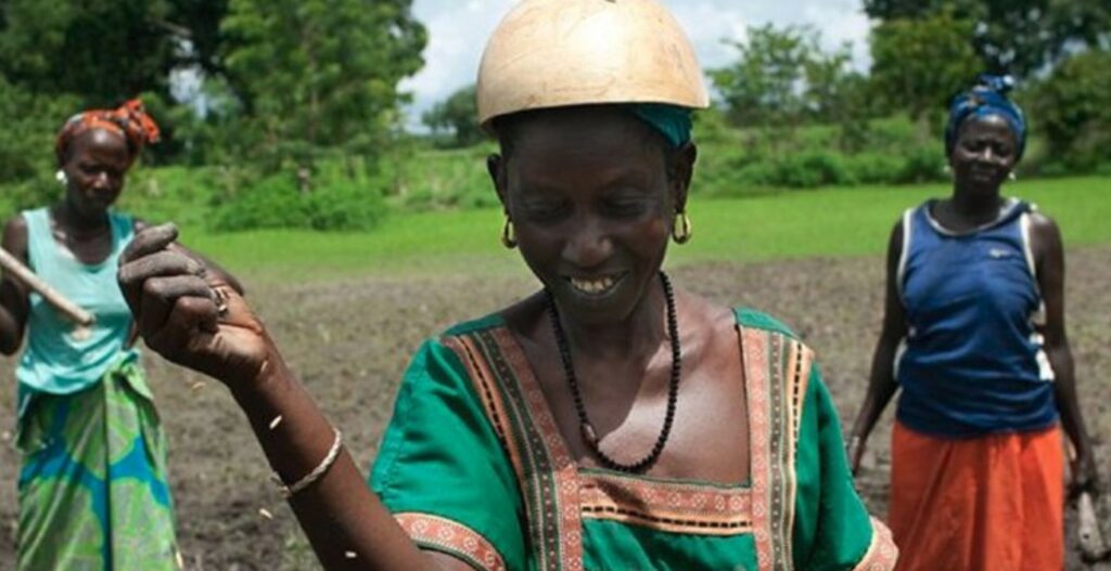 A woman with a helmet smiling and holding a hoe, with two women in colorful skirts and tops working in a green field behind her. The scene depicts community agricultural work in a rural setting.