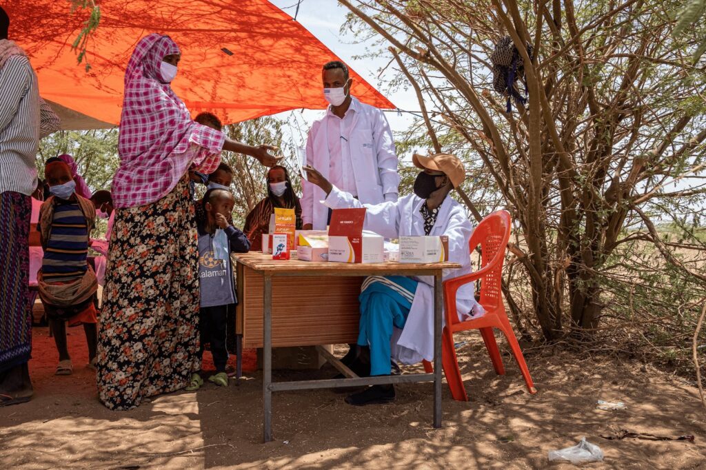 Medics distributing medication to children in a refugee camp, under a makeshift shade with aid supplies on a table, people lining up for health services in a rural area, community health outreach effort for displaced populations.