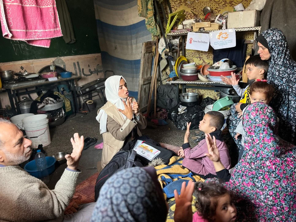 Fearless women and children gather in a modest home, engaging in heartfelt discussions amid basic kitchen supplies, highlighting the urgent need for humanitarian aid and community support.