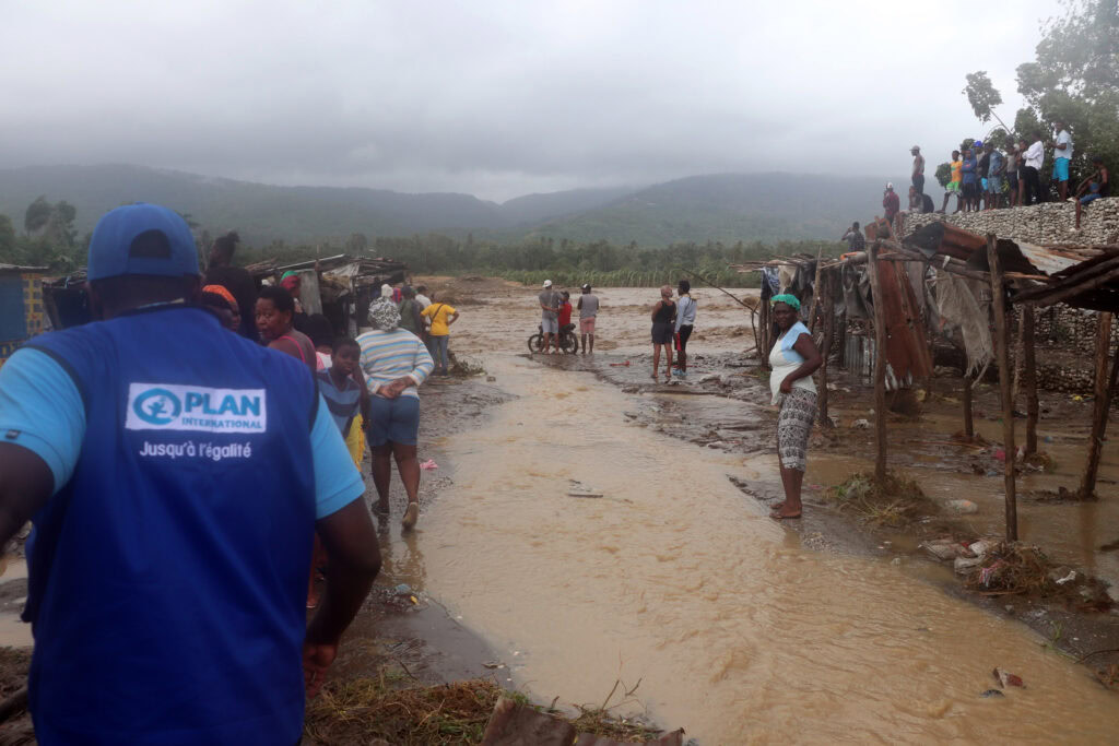 Flooded area with people assessing damage and providing aid in a rural setting under cloudy skies. Local communities and aid workers are present amidst destroyed structures and muddy water, highlighting disaster response efforts.
