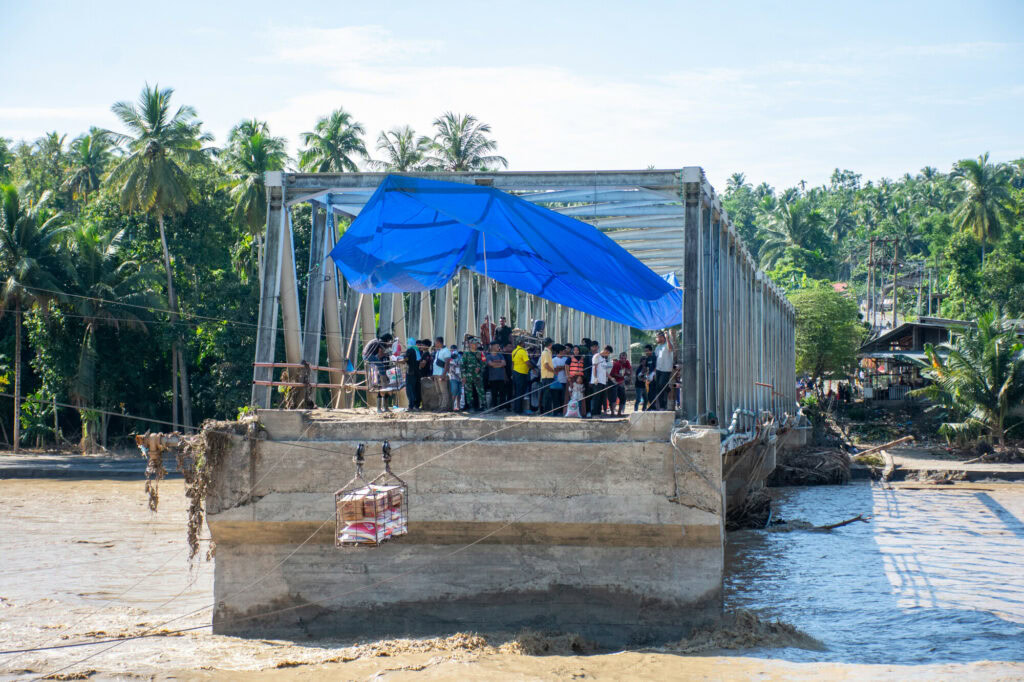 People gather on a partially constructed bridge with a blue tarp for shade, amidst tropical trees and a river, illustrating emergency aid efforts by the Dutch Relief Alliance in a crisis situation.