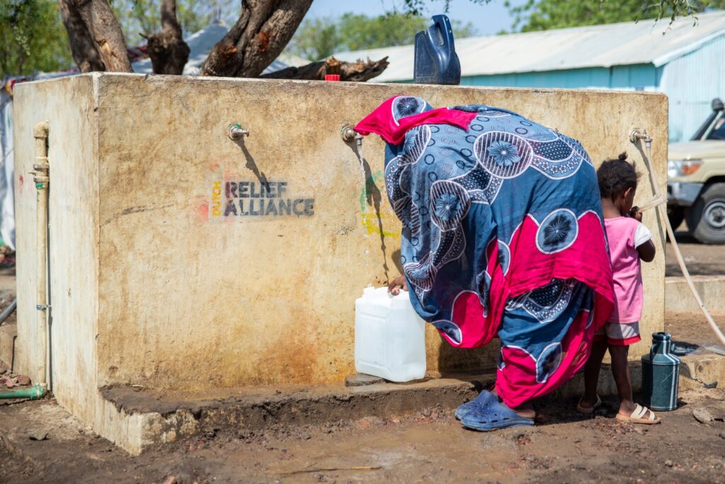 1. Woman filling a water container from an emergency water station marked with "RELIEF ALLIANCE" in a rural area.