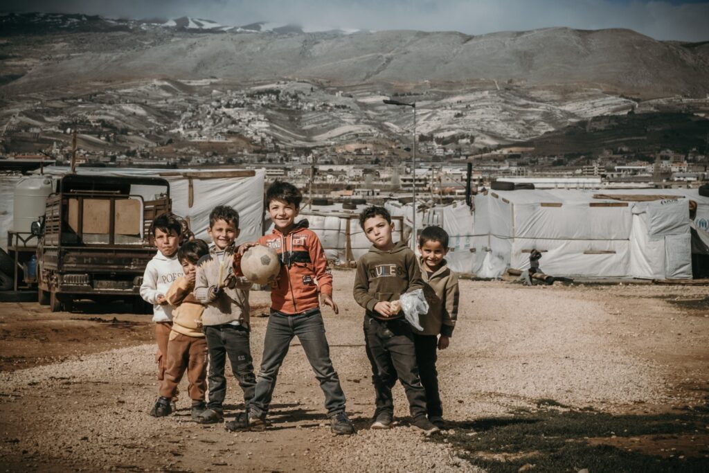 Children playing outdoors in a refugee camp with tents and mountains in the background.
