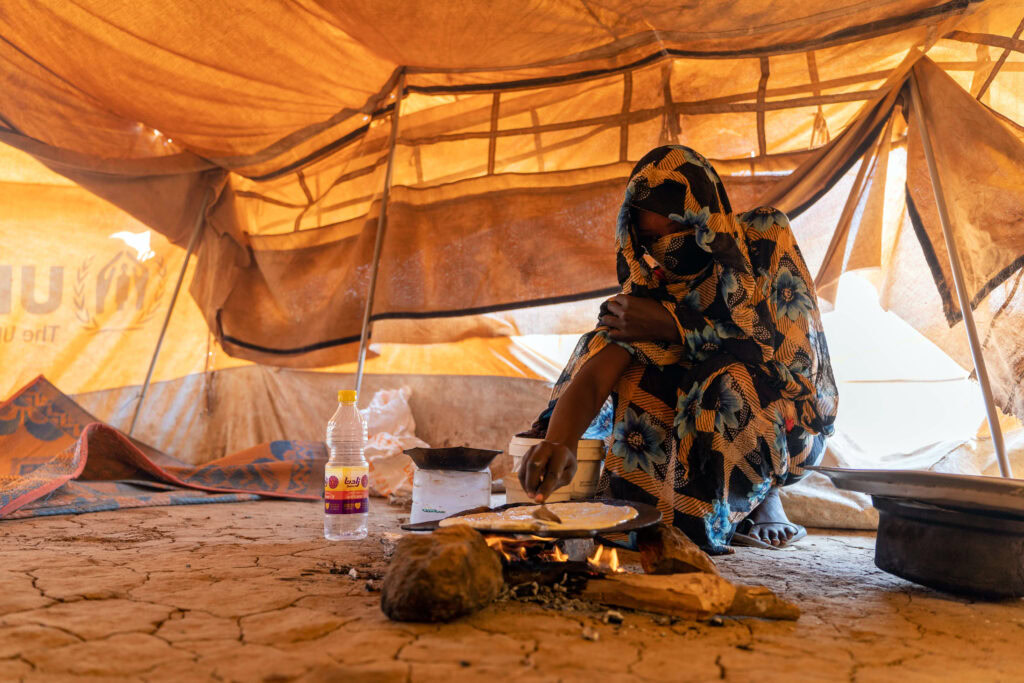 Woman in traditional clothing sitting inside a tent, preparing food or supplies amidst a humanitarian emergency.