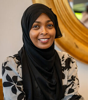 Een vrouw met een zwarte hijab en gedessineerde blouse, binnen zittend met een warme glimlach, voor een grote ronde spiegel, met vage mensen op de achtergrond.