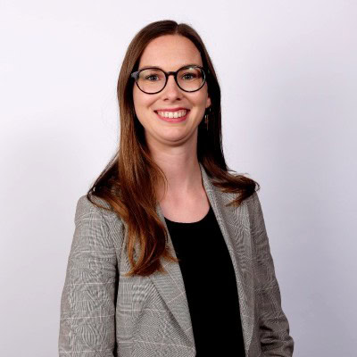 Een professionele vrouw met bril, glimlachend, met een grijze blazer en zwart topje, staand tegen een effen lichte achtergrond.