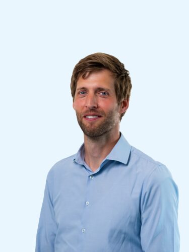 Een professioneel portret van een man in een lichtblauw overhemd, zelfverzekerd glimlachend tegen een effen lichte achtergrond, die staat voor effectieve communicatie en paraatheid bij noodgevallen.