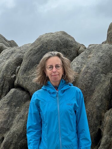 Vrouw met blauwe jas staat buiten tussen grote rotsen, met een bewolkte lucht boven haar hoofd.