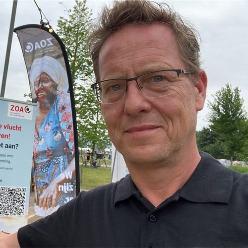 Een man met bril en krullend haar staat buiten op een hulpverleningsevenement en houdt een bord vast met informatie over noodhulp en contactgegevens, met een vlag en bomen op de achtergrond.