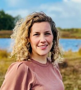 Een glimlachende vrouw met krullend blond haar staat in een natuurlijk landschap met water en groen onder een blauwe lucht.