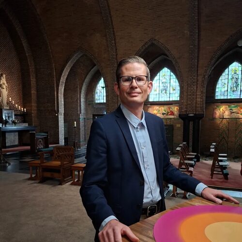 Een professionele man in pak staat in een kerk met gebrandschilderde ramen, houten kerkbanken en bakstenen bogen, wat een formele of religieuze omgeving laat zien.