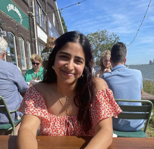 Een jonge vrouw met lang donker haar en een gebloemde rode jurk geniet van een zonnige dag in een openluchtcafé aan het water, omringd door andere klanten en een schilderachtig uitzicht.