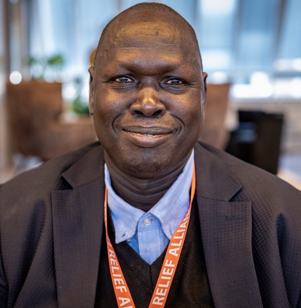 Een portret van een lachende man in een pak met een koordje met "Dutch Relief Alliance" erop, in een interieur met onscherpe achtergrond.