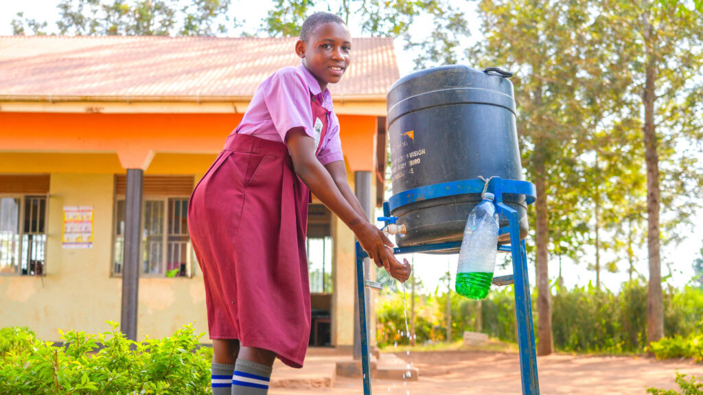 Young girl in school uniform using a handwashing station outdoors in a rural setting.
