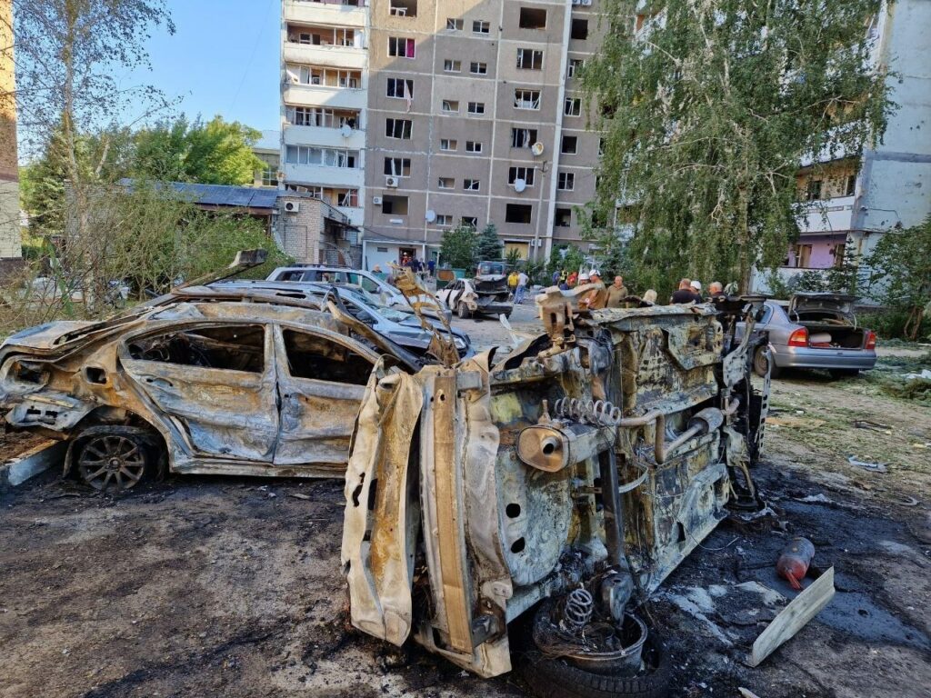 Burned-out vehicle and damaged buildings in an urban area after a fire or explosion.
